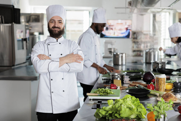Head chef wearing cooking uniform while standing in restaurant professional kitchen with arms crossed. Gastronomy expert cutting fresh vegetables and mixing spices for dinner service. - Photo, Image