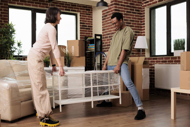 Diverse couple unpacking furniture to decorate new real estate flat bought on mortgage loan, enjoying household relocation after moving in together. New beginnings with apartment property. - Photo, Image