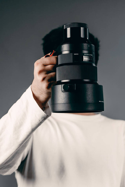 A vertical shot of a man holding a black professional lens in front of his face - Foto, Imagen