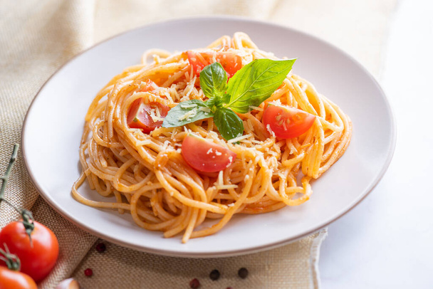 Tasty appetizing classic italian spaghetti pasta with tomato sauce, cheese parmesan and basil on plate and ingredients for cooking pasta on white marble table. - Foto, Imagen