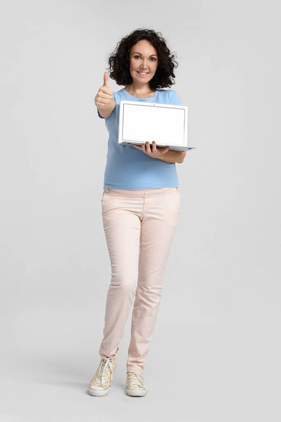 Młoda kobieta w ciąży z laptopem pokazującym kciuk do góry na jasnym tle - Zdjęcie, obraz