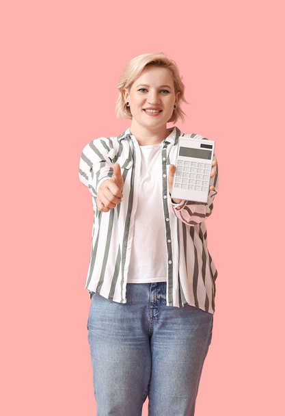 Schöne blonde Frau mit Taschenrechner zeigt Daumen nach oben auf rosa Hintergrund - Foto, Bild