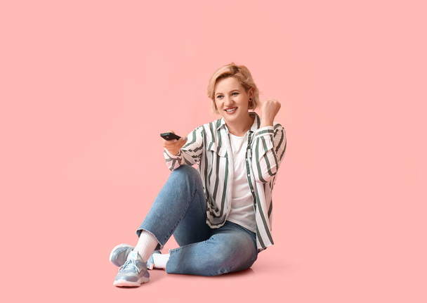 Beautiful blonde woman with TV remote control sitting on pink background - Фото, изображение