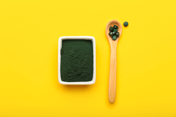 Bowl of spirulina powder and spoon with pills on yellow background - Photo, Image