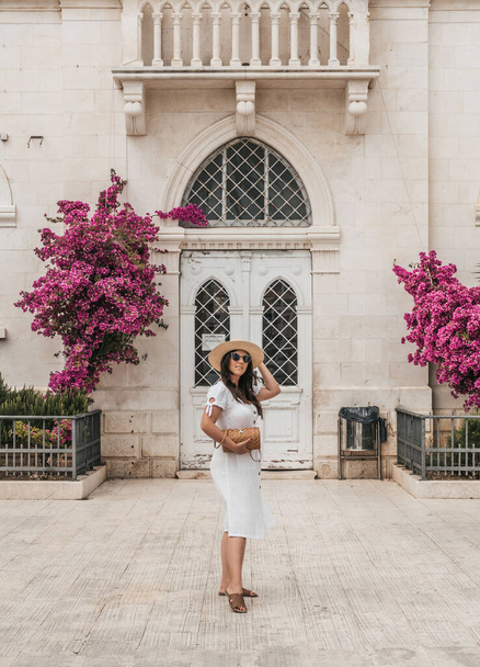 A vertical shot of a Caucasian woman in a white dress posing in front of an old building with bougainvillea flowers growing outdoors. - Photo, image