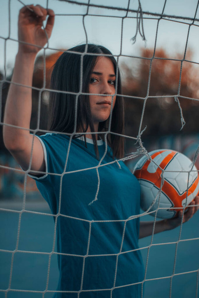 A vertical shot of a beautiful female football player posing behind the football net - 写真・画像