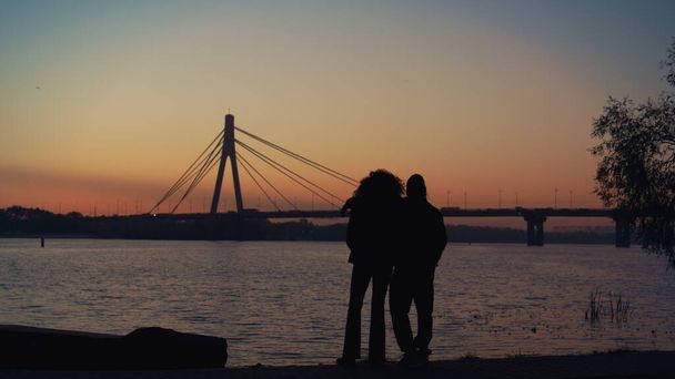 Liebhaber beobachten den Sonnenuntergang am Ufer des Flusses. Schöner klarer Abendhimmel. Unbekanntes Paar umarmt Brücke noch nachts Wasserstraße genießen romantisches Date. Emotionales Verbindungskonzept. - Foto, Bild