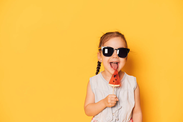 Girl in glasses licks lollipop in the form of piece of watermelon against yellow background. - Photo, Image