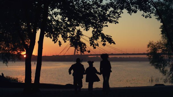 Gelukkige familie silhouet lopen naar de rivier hand vasthouden. Prachtig uitzicht op de brug. Onbekende liefhebbende ouders genieten van opvoeding veel plezier met kind avond hemel. Emotionele verbinding zorgeloos kinderconcept - Foto, afbeelding