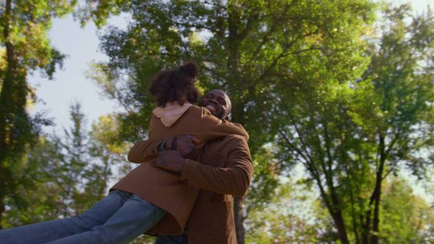 Ευγενικός πατέρας αγκαλιάζει κόρη γυρίζοντας παιδί στο ηλιόλουστο πάρκο. Αγαπημένες γονικές αγκαλιές. Χαρούμενος Αφροαμερικάνος χαμογελαστός μπαμπάς που απολαμβάνει να εκφράζει την αγάπη του. Συναισθηματική σύνδεση ανέμελη παιδική ηλικία έννοια. - Φωτογραφία, εικόνα