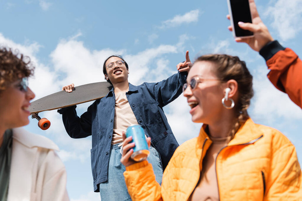 lage hoek uitzicht van aziatische man met longboard tonen overwinning teken in de buurt van gelukkige vrienden - Foto, afbeelding