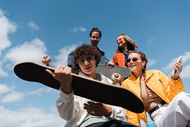 laag gezichtspunt van jonge man houden skateboard in de buurt gelukkig interraciale vrienden buiten - Foto, afbeelding
