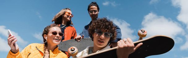 vista de ángulo bajo de los patinadores interracial cerca de amigos alegres contra el cielo azul nublado, pancarta - Foto, Imagen
