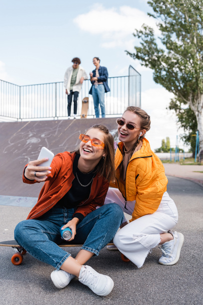 radostné ženy při selfie v skate parku v blízkosti přátel na rampě na rozmazaném pozadí - Fotografie, Obrázek