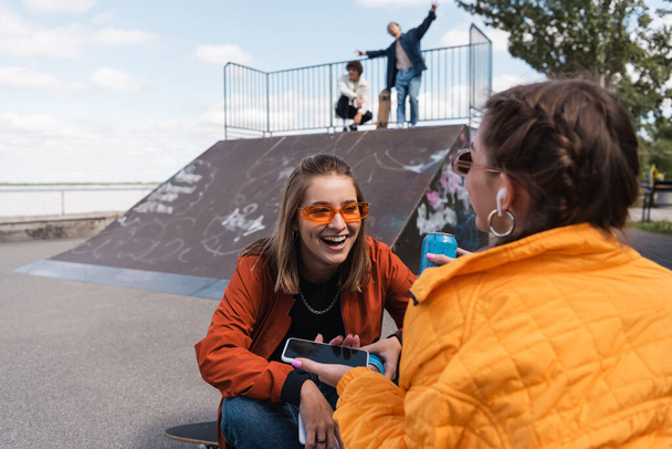 mujer sonriente en gafas de sol con estilo sentado cerca de amigo y los hombres borrosos en la rampa de skate - Foto, imagen