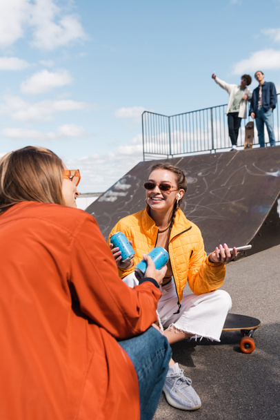uśmiechnięta kobieta w okularach rozmawia ze znajomym w pobliżu rozmytych łyżwiarzy na rampie w skate parku - Zdjęcie, obraz