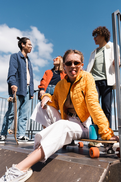 женщина в солнцезащитных очках высовывает язык, сидя на скейтборде рядом с межрасовыми друзьями - Фото, изображение