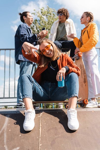 trendy woman with soda can showing victory sign while sitting on skate ramp near blurred interracial friends - Photo, Image