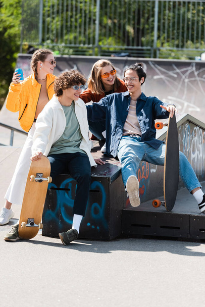 cheerful and trendy multiethnic friends sitting on ledge in skate park - Photo, Image