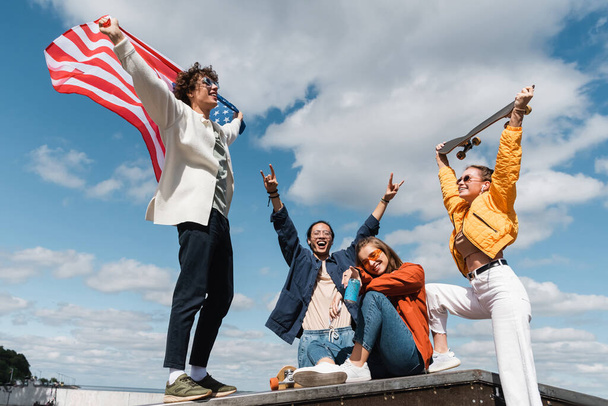emocionado asiático hombre mostrando rock signos cerca de amigos con EE.UU. bandera y monopatín - Foto, Imagen