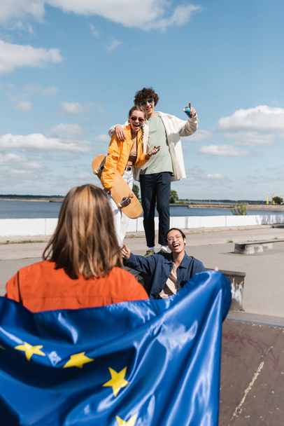 heureux amis multiculturels pointant vers la femme avec le drapeau de l'union européenne sur le premier plan flou - Photo, image