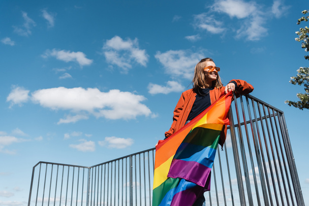 low angle view of smiling woman with lgbt flag against blue cloudy sky - Zdjęcie, obraz