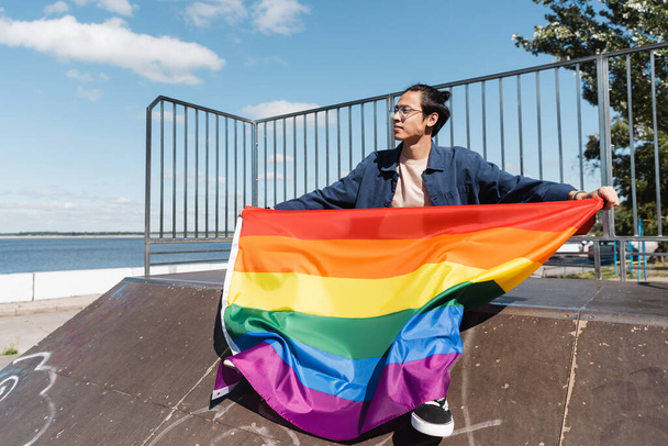 молодой азиат с флагом гордости сидит на рампе скейт-парка - Фото, изображение