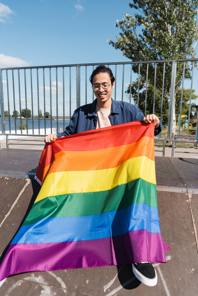 счастливый азиатский мужчина в очках сидит на рампе в скейт-парке с флагом - Фото, изображение