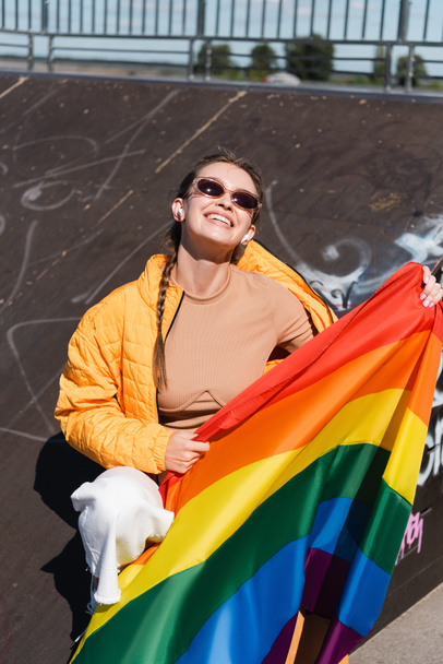 happy woman in wireless earphones sitting with lgbt flag on ramp in skate park - Фото, изображение