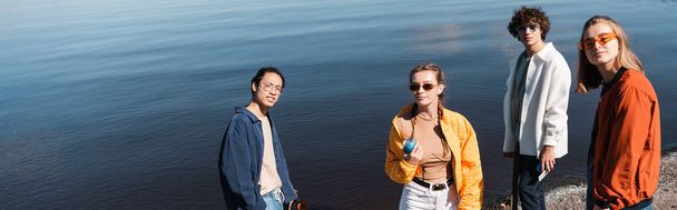 young woman holding soda can near multicultural friends and river, banner - 写真・画像