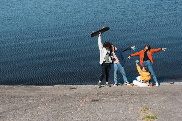cheerful multiethnic skaters having fun on embankment near river - 写真・画像