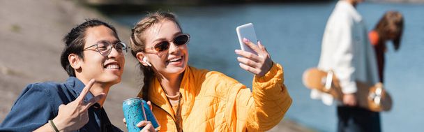 glückliche Frau mit Sonnenbrille macht Selfie mit asiatischem Freund und zeigt Siegesgeste, Banner - Foto, Bild