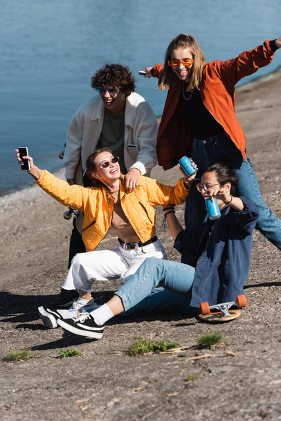 mujer feliz con teléfono inteligente y refresco puede divertirse con amigos interracial en terraplén - Foto, Imagen