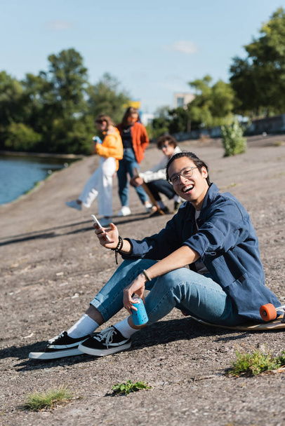 szczęśliwy azjatycki łyżwiarz z puszką sody i telefon komórkowy siedzi na nasypie w pobliżu rozmazanych przyjaciół - Zdjęcie, obraz