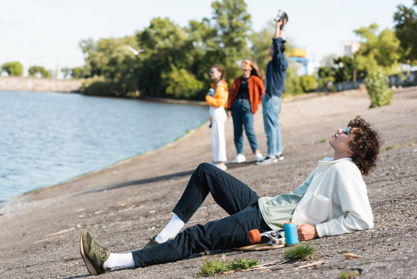 full length of young skater relaxing on embankment near blurred friends - Photo, Image