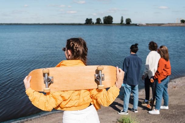 back view of young woman with skateboard standing near river and blurred friends - Foto, afbeelding