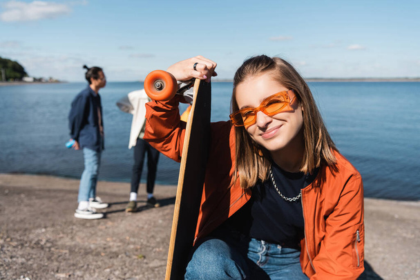 happy and stylish woman with longboard looking at camera near blurred friends on riverside - 写真・画像