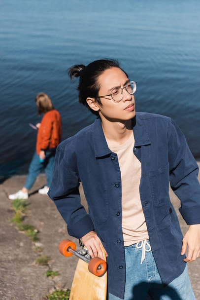 young asian skater in blue shirt and eyeglasses looking away on riverside - Fotografie, Obrázek