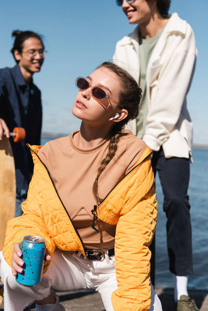 young woman in earphone and sunglasses holding soda can near blurred interracial friends - Foto, imagen