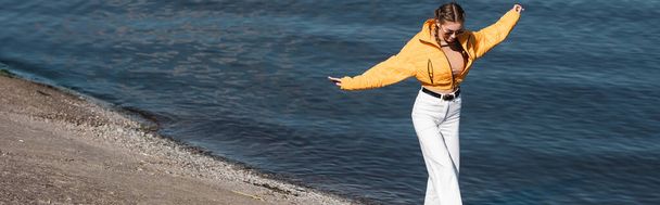 mujer en chaqueta amarilla caminando con las manos extendidas a lo largo de la orilla del río, bandera - Foto, imagen