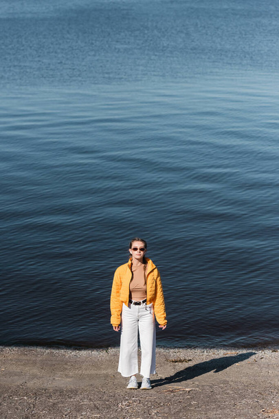 longitud completa de la mujer en gafas de sol y ropa de moda de pie cerca del río - Foto, imagen