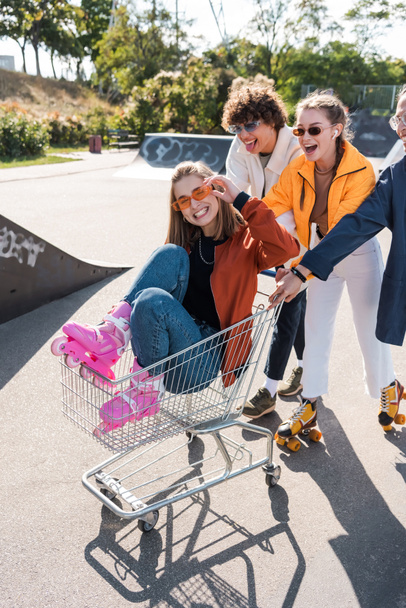 smiling woman adjusting sunglasses while riding in shopping cart near friends - Photo, Image