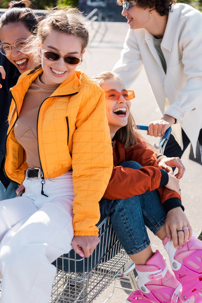 Ευτυχισμένοι πολυπολιτισμικοί φίλοι με γυαλιά ηλίου διασκεδάζοντας με το καροτσάκι αγορών σε εξωτερικούς χώρους - Φωτογραφία, εικόνα