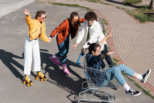 cheerful women holding hands and riding roller skates near asian man in shopping trolley - Fotografie, Obrázek