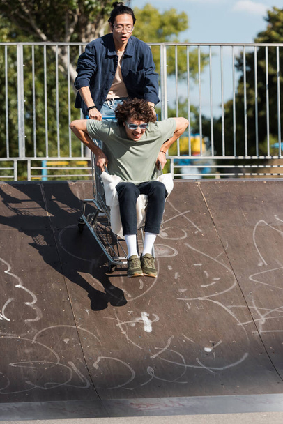 cheerful interracial friends having fun with shopping cart on ramp in skate park - Photo, Image