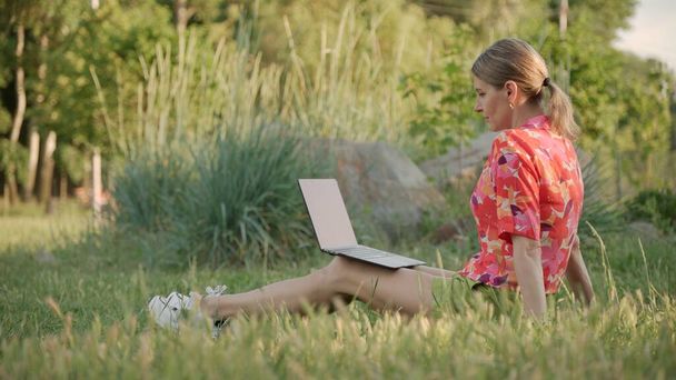 Young woman sits on grass in park and looks at screen of her laptop. Distance learning. - Photo, Image
