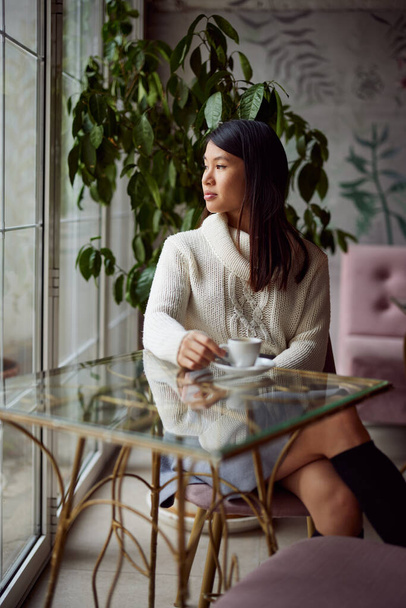 A thoughtful asian woman is sitting in a coffee shop and looking trough the window while drinking a coffee. - Foto, afbeelding