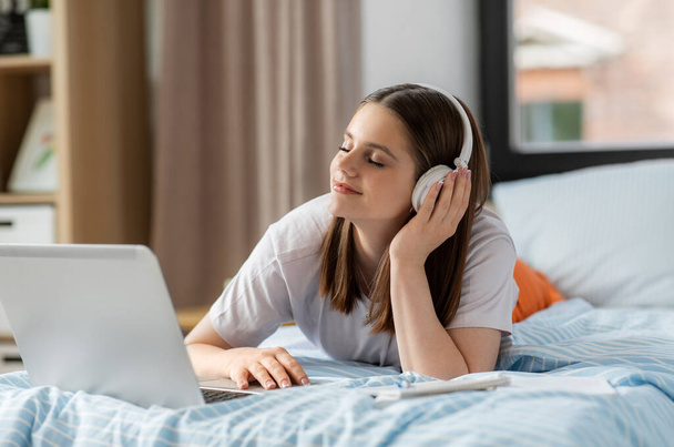 school, online education and e-learning concept - happy smiling teenage student girl in headphones with laptop computer lying on bed and listening to music at home - Fotó, kép