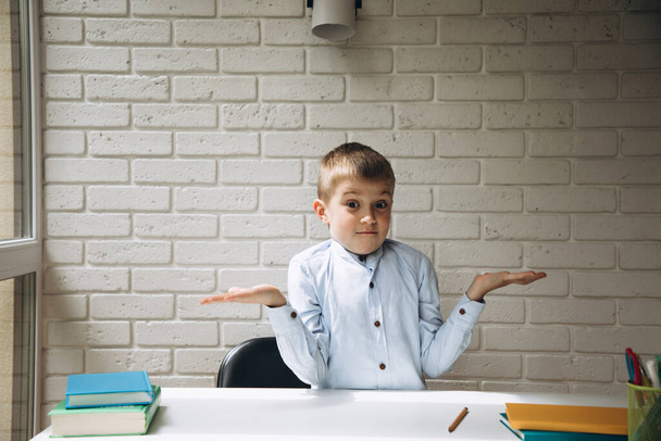 Chłopiec w wieku 6-7 lat przy białym biurku pokazuje nieznany gest przeciwko ceglanej ścianie. Edukacja szkolna - Zdjęcie, obraz