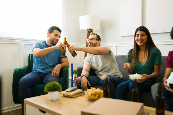 Hispanische Freunde klirren mit ihren Bierflaschen, während sie vor dem Fernseher sitzen und das Sportspiel genießen  - Foto, Bild